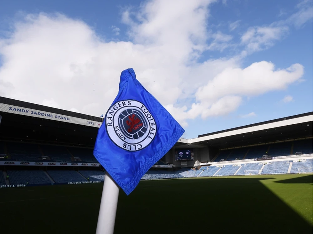 Eine blaue Eckfahne mit dem traditionellen Wappen des Rangers Football Club vor den leeren Tribünen der Sandy Jardine Stand unter blauem Himmel.